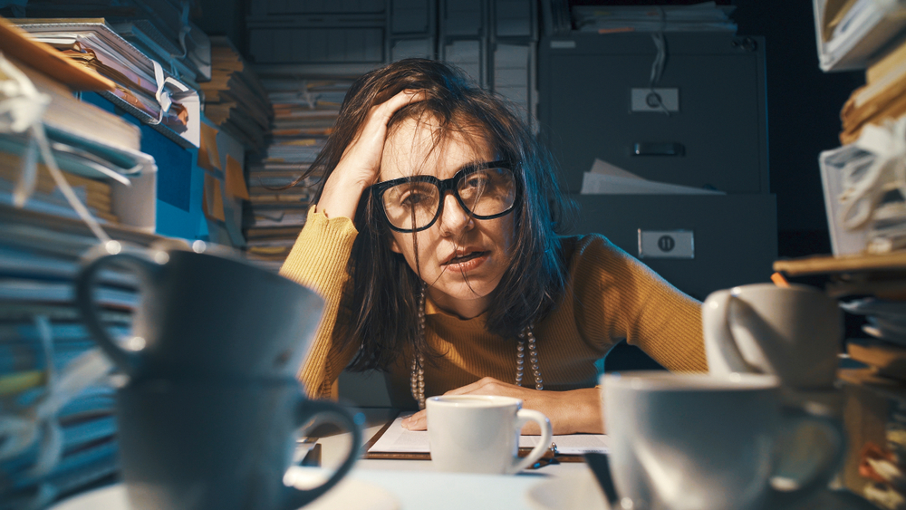 Young woman at her desk, running her fingers through her hair as she contemplates her workload.  Surrounded by books and papers, empty coffee cups stacked on the desk