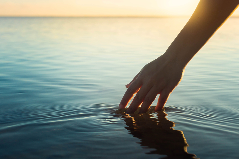 Serene scene of arm and hand with fingertips dipping into a placid body of water at sunset