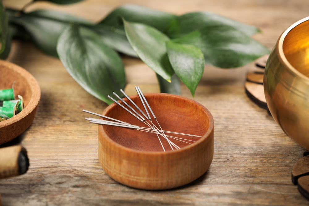 Serene setting with wooden bowl containing an array of acupuncture needles.  Leafy background.  Moxabustion stick in foreground.